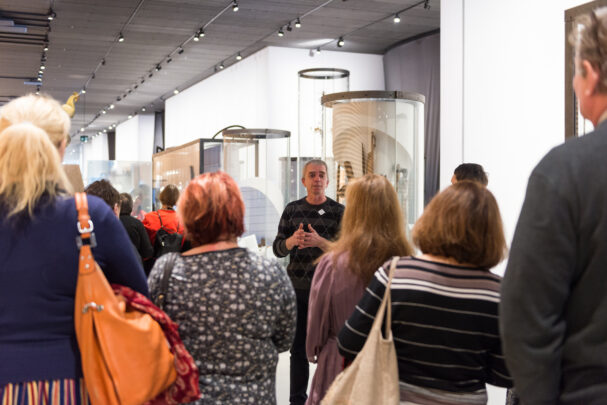 Rääkiv mees seisab näitusesaalis publiku ees ning grupp kuulajaid seisab tema ümber, vaadates ja kuulates eksponeeritud esemeid.