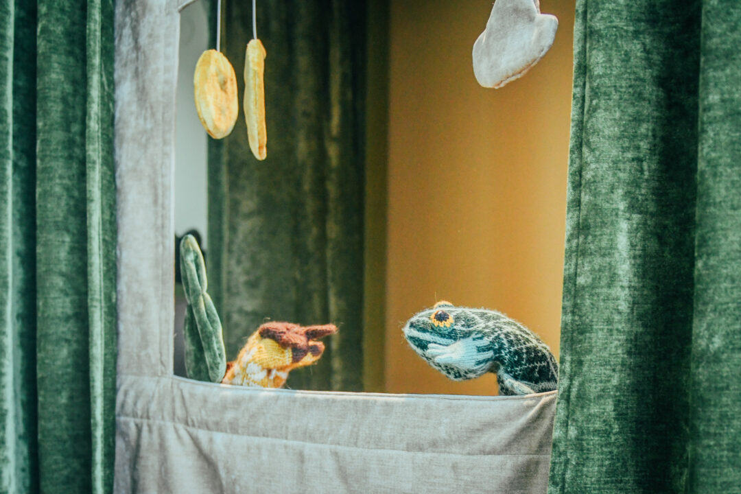 Roheliste sametkardinatega aknast vaatavad välja kaks käpiknuku looma, üks sinakas-pruun ja teine kollakas-pruun.